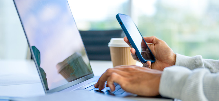 Decorative image. A person on a laptop with a mobile in their hand and a cup of coffee